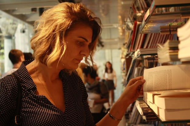 A person browsing a selection of books in a bookstore, with a focus on the spines of books by diverse authors. The bookstore is well-lit and inviting, with other customers browsing in the background.