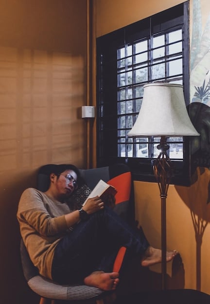 A relaxed photo of the author being interviewed. The author is sitting in a comfortable chair, holding a copy of one of their novels, and smiling warmly at the interviewer, who is partially visible on the side. The setting is a cozy living room, with bookshelves in the background.