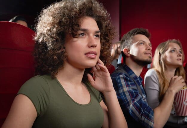 A close-up of a theater critic in the audience, intently watching the stage. She is holding a notepad and pen, seemingly capturing notes on the performance. The background is slightly blurred, focusing the viewer's attention on her focused expression.