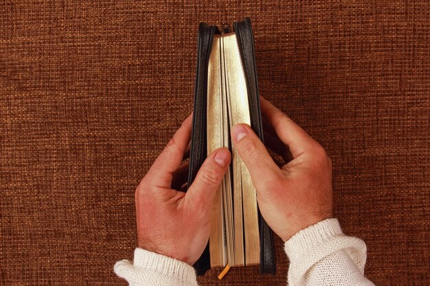 A close-up shot of a person's hands holding an open book, with their fingers gently turning the page. The book is set in a cozy, dimly lit environment. The focus is on the tactile sense of reading and how personal it can be. A cup of tea/coffee is blurred in the background.